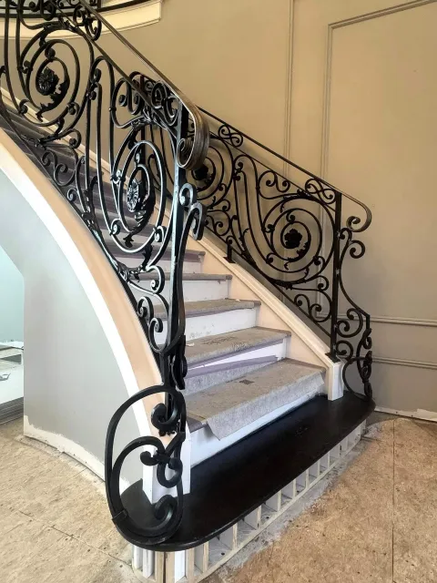 Interior view of a staircase with intricately designed black wrought iron railing featuring swirling patterns and floral details. The stairs are made of light-colored materials, possibly marble or granite, with visible steps and a black platform at t