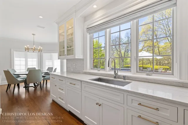 Bright kitchen with large window overlooking trees, white cabinetry, marble countertops, stainless steel sink, and a dining area with a round table and gray chairs.