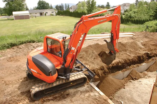 Orange excavator digging a trench in a dirt field near a house