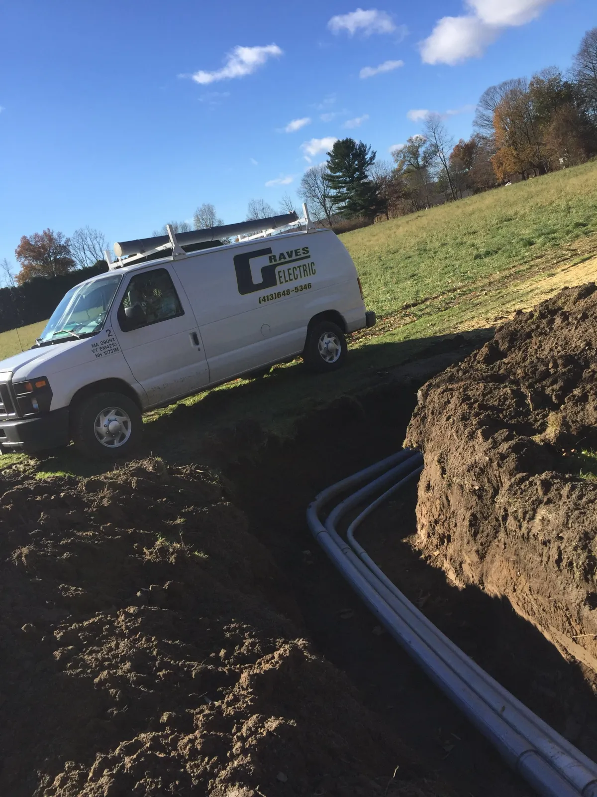 A white van with the company name Raves Electric parked on a grassy field next to an open trench with electrical conduits laid inside. The scene is outdoors on a sunny day with a clear blue sky and some scattered clouds.
