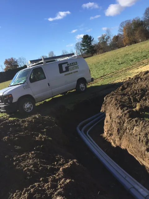 A white van with the company name Raves Electric parked on a grassy field next to an open trench with electrical conduits laid inside. The scene is outdoors on a sunny day with a clear blue sky and some scattered clouds.