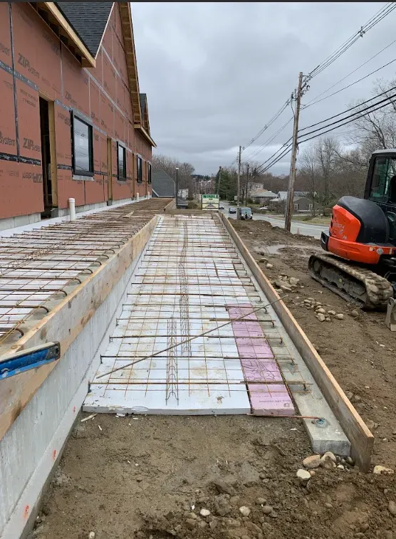 Construction site with a narrow concrete walkway beside a brick building and an excavator on the right.