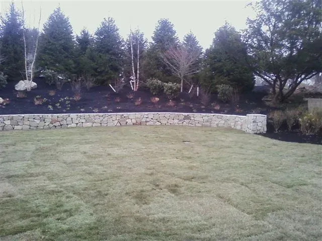 Curved stone retaining wall beside a grassy lawn, with trees and shrubs along the slope.