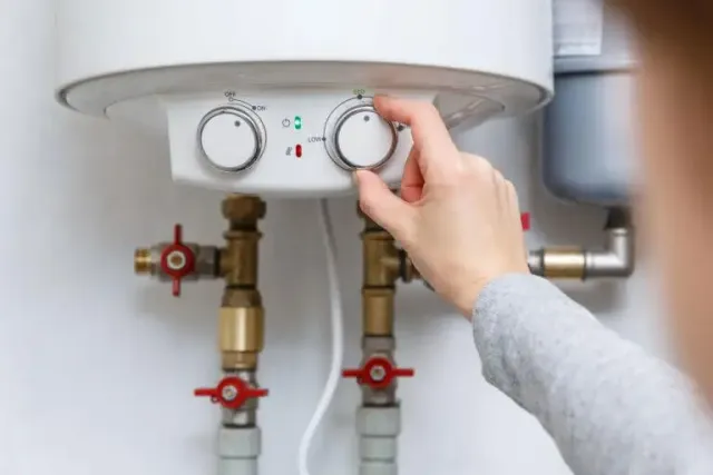 Person adjusts the temperature dial on a wall-mounted water heater, with red valve handles and brass fittings visible below