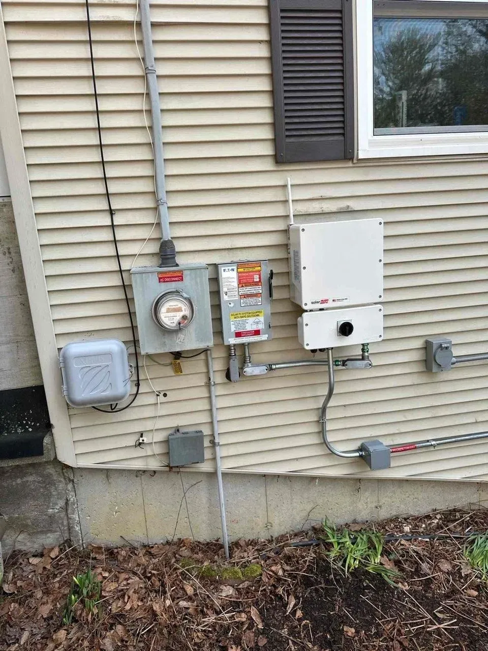 Outdoor house utility meters and electrical boxes mounted on beige siding beside a window.