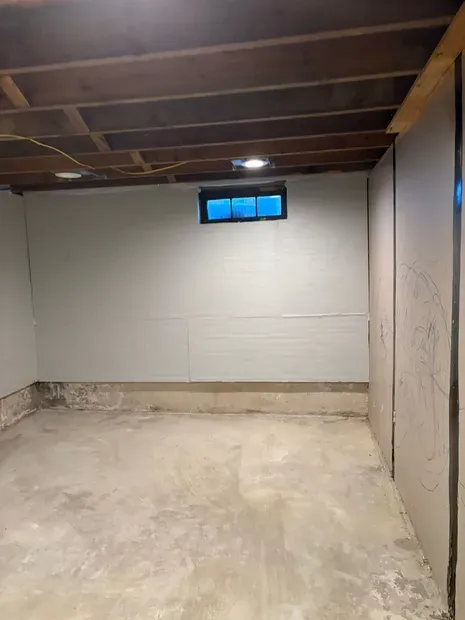 Empty basement room with unfinished walls, exposed ceiling joists, and a small high window.