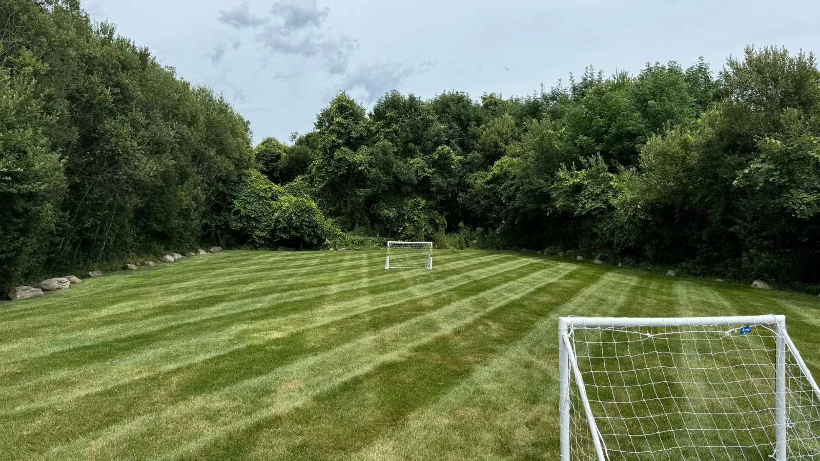 Soccer goals on a striped, green lawn surrounded by trees under a cloudy sky.