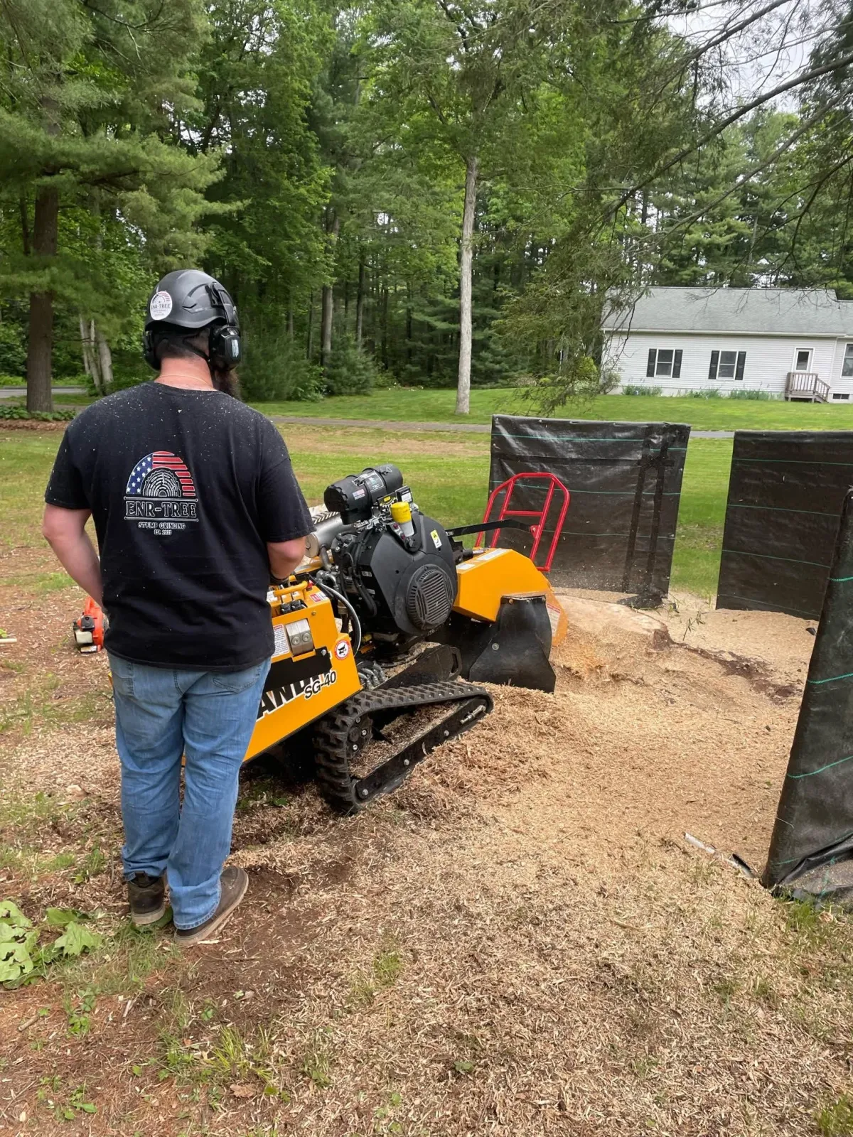 A person wearing a protective helmet and a T-shirt with an American flag fingerprint design stands near a compact skid-steer loader in a backyard with green trees and a white house in the background.