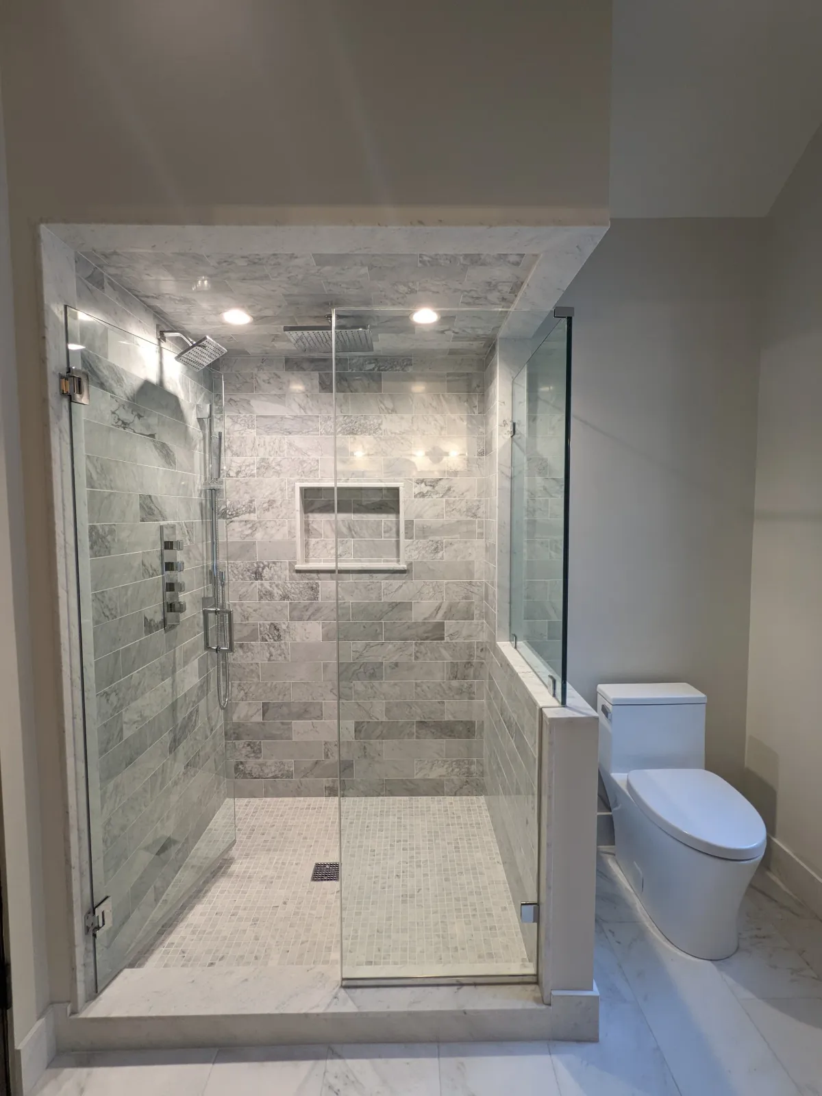 Modern bathroom with a glass-enclosed shower featuring grey marble tiles, a built-in shelf, and a rainfall showerhead, next to a white toilet.