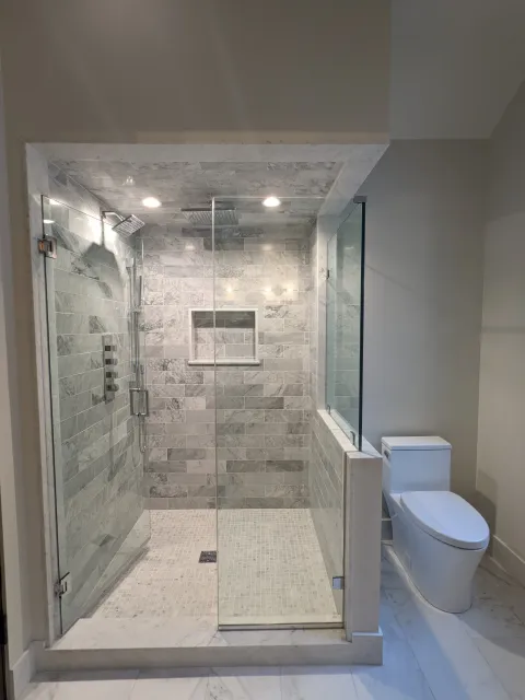Modern bathroom with a glass-enclosed shower featuring grey marble tiles, a built-in shelf, and a rainfall showerhead, next to a white toilet.