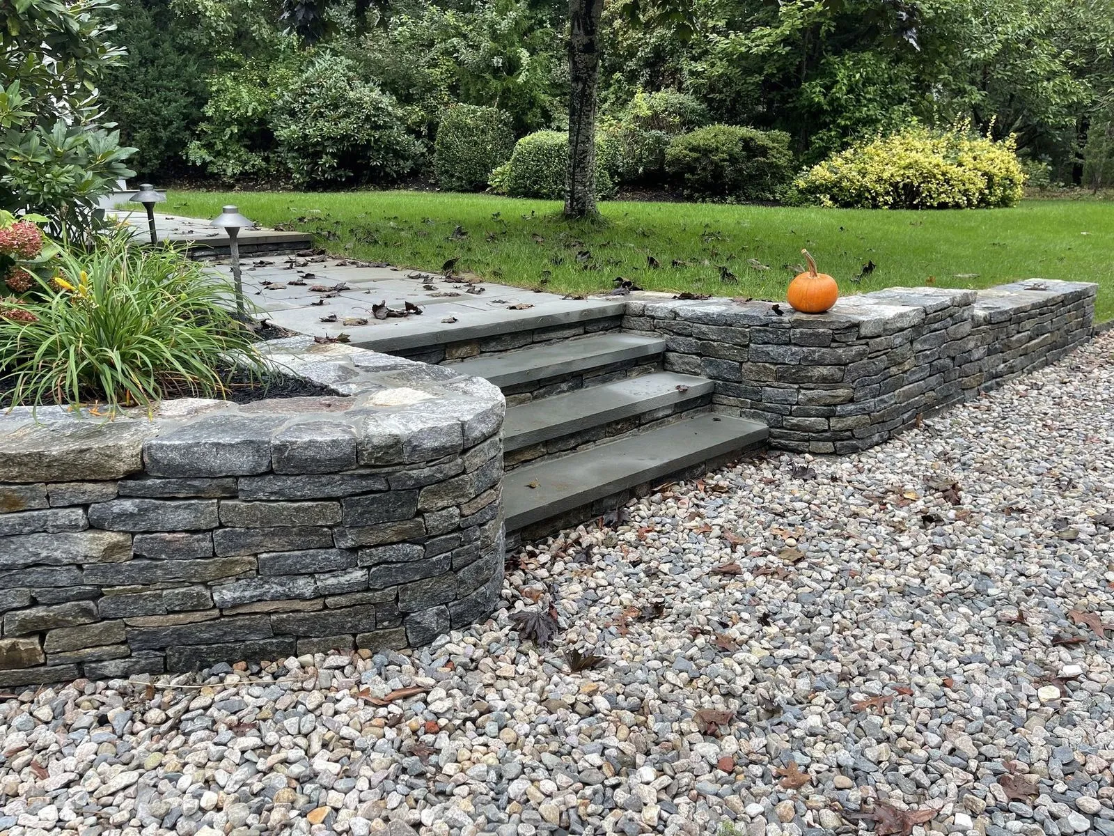 Bluestone steps set between drystack stone retaining walls, river-rock drainage at base, autumn pumpkin on the cap stone
