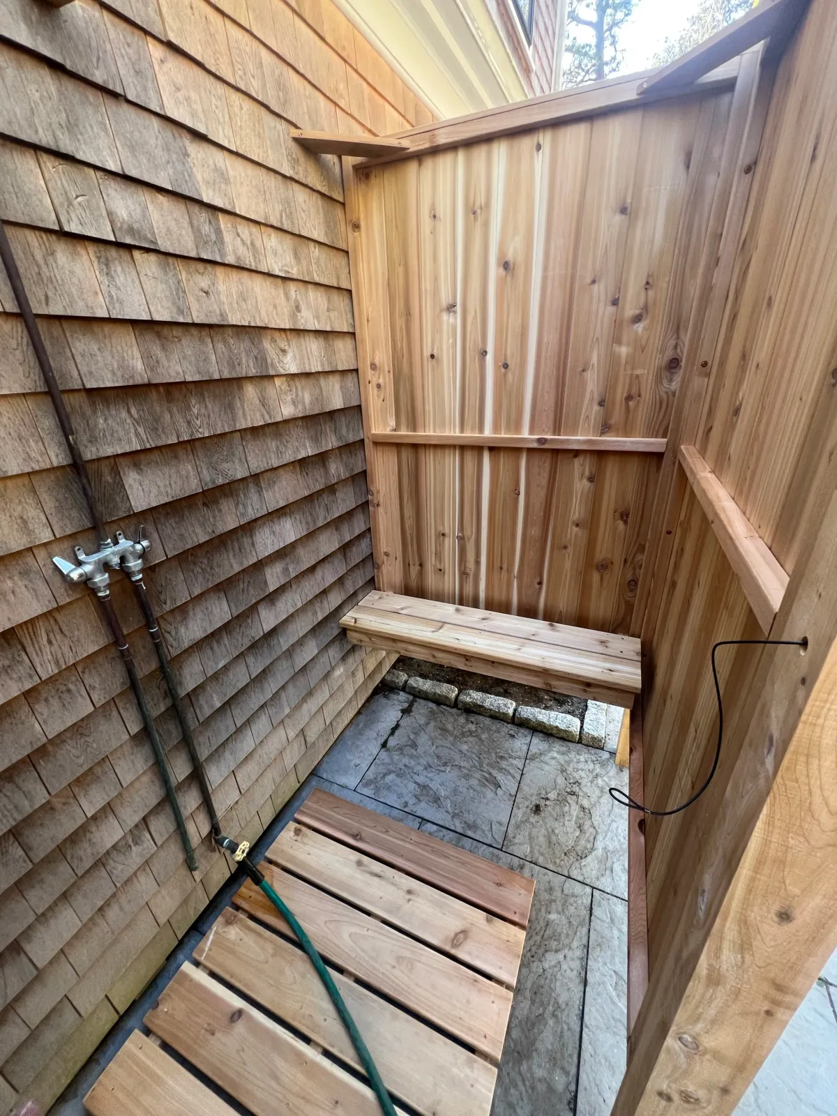 A small outdoor shower area with wooden walls, a stone tile floor, and a bench, with stored water pipes visible on the left side.