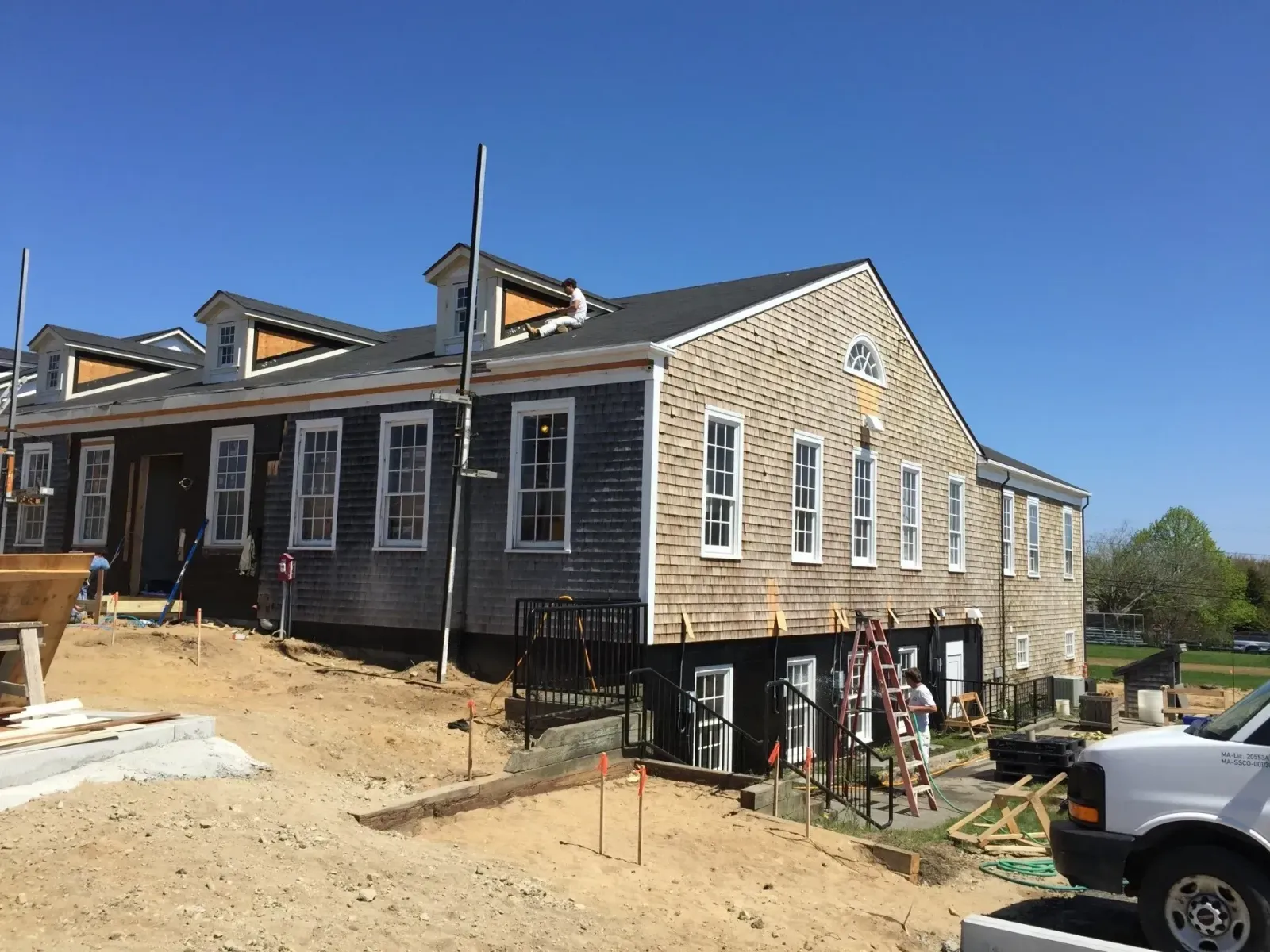 Building under construction, weathered gray siding, white trim, blue sky. Construction workers on site.