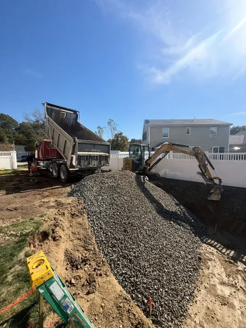 Truck dumping gravel into an excavator, building a retaining wall on a sunny day.