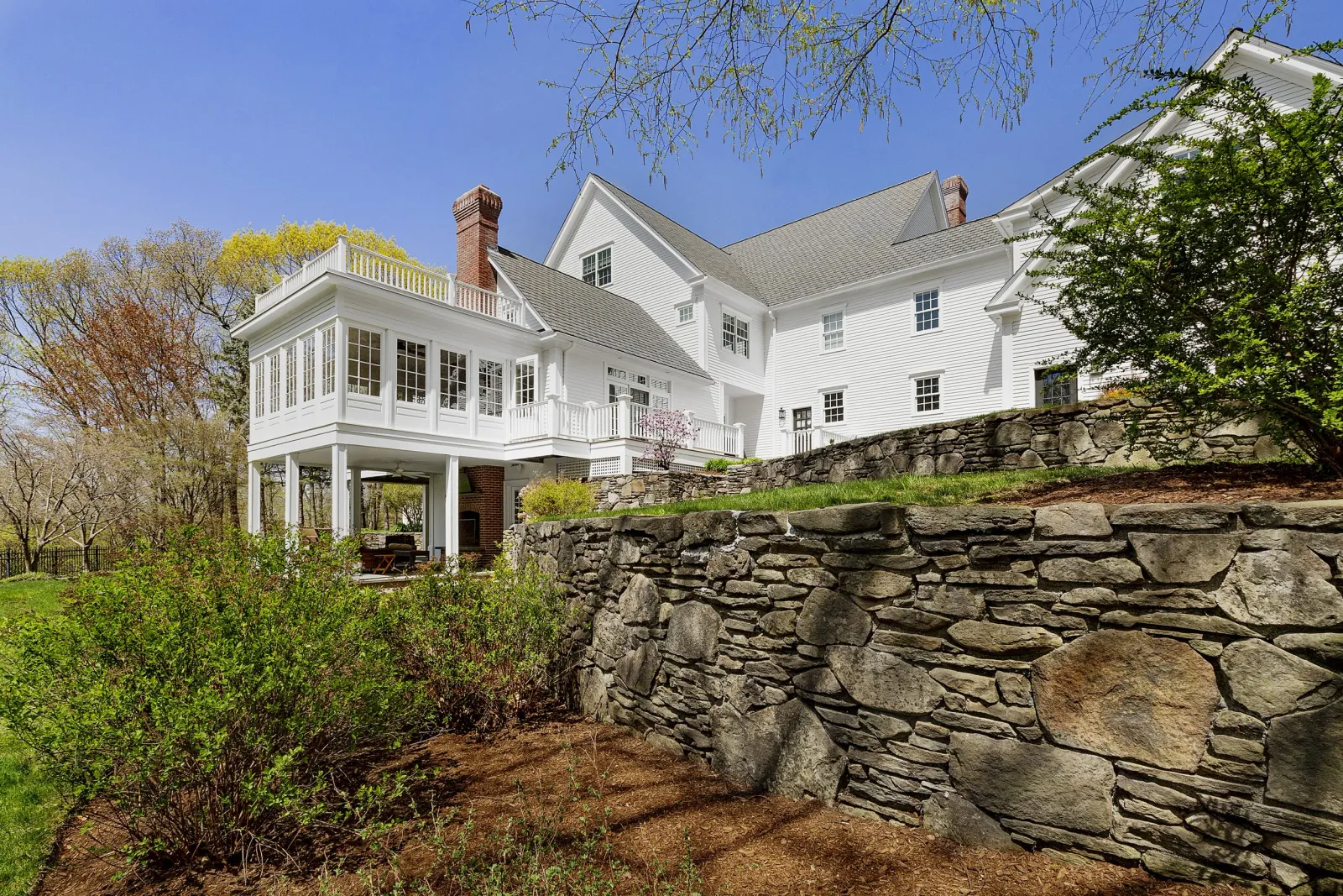 Large white house with multiple windows and brick chimneys, surrounded by trees and a stone retaining wall.