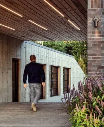 A man in casual clothes walks toward a modern building with wooden and brick elements, designed by a Maine architect, surrounded by plants and trees.