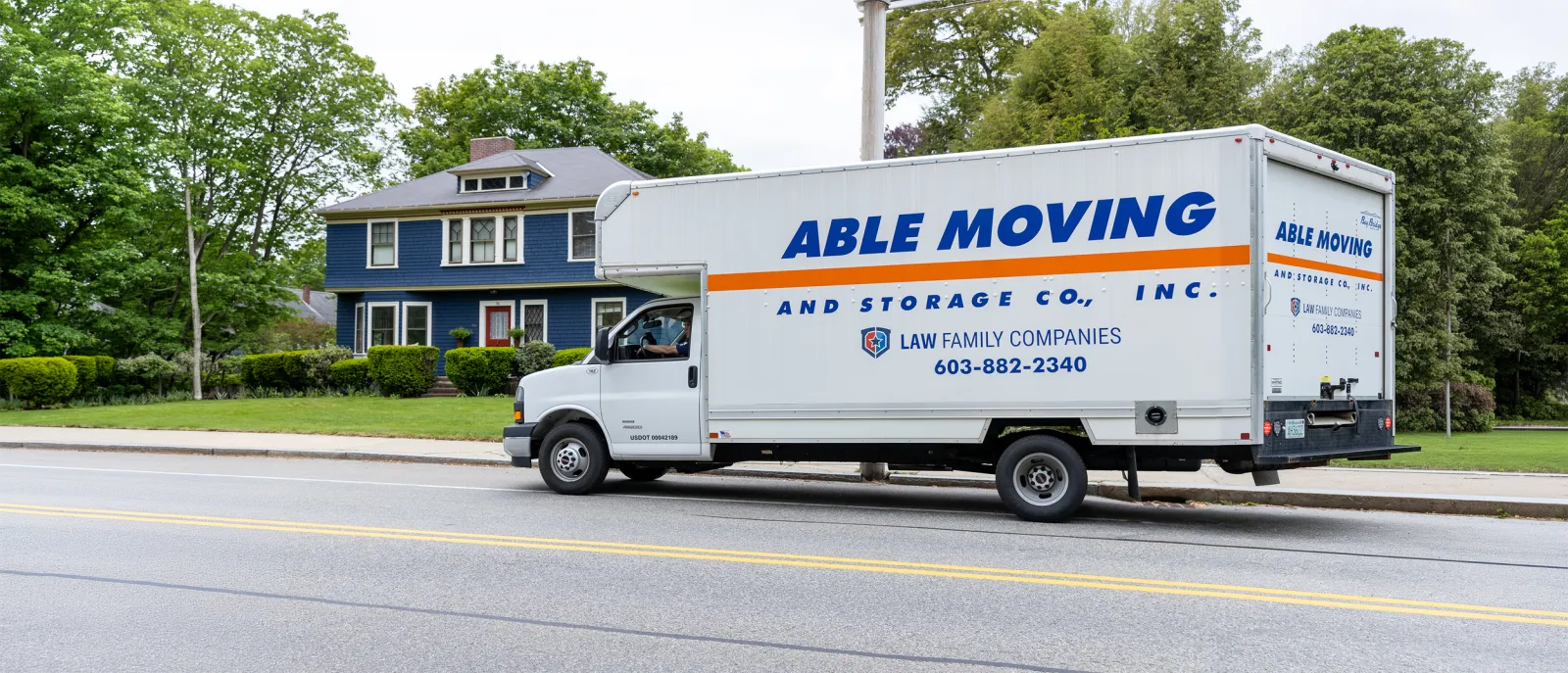 A white moving truck with blue and orange lettering reading 'Able Moving and Storage Co., Inc.' parked on a suburban street in front of a large blue house with green trees in the background.