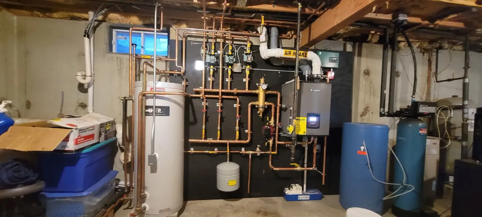 A utility room basement setup featuring a grey boiler, copper piping, a water heater, and water filtration equipment.
