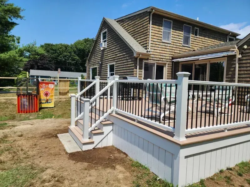 Raised wooden deck with white railings beside a house, in a sunny backyard
