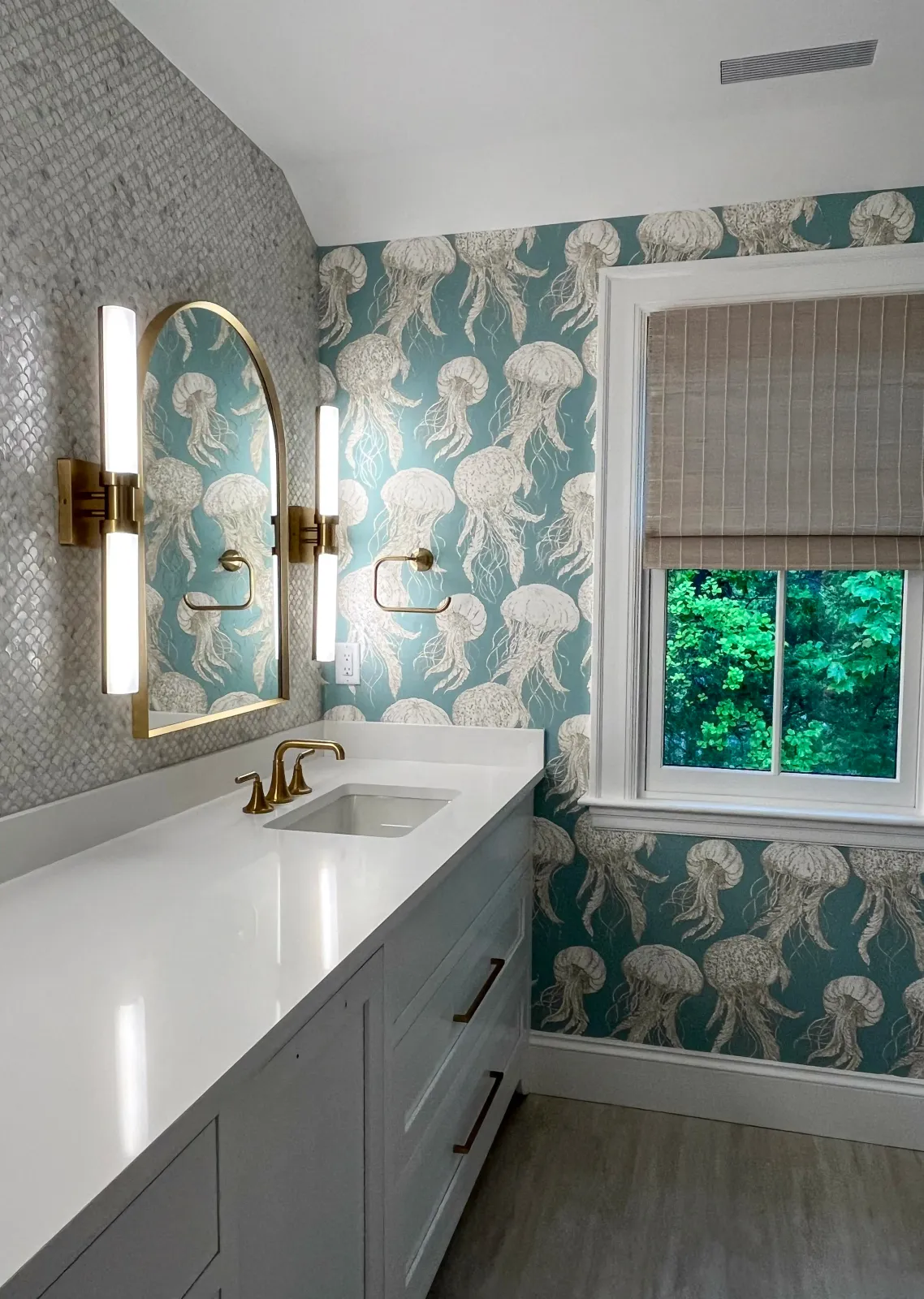 Bathroom vanity with white countertop, gold fixtures, and a jellyfish pattern wallpaper underneath a window with a beige blind.
