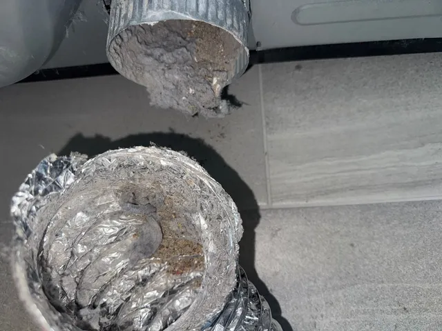 A section of dryer duct laid on the floor with its interior lined in compacted lint and grey debris.