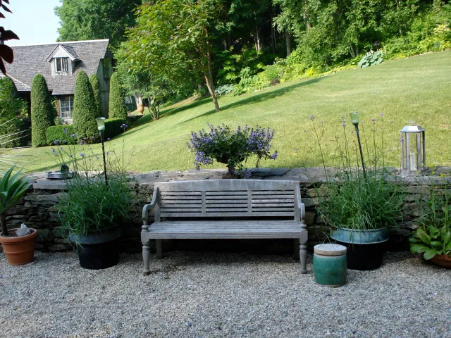 A backyard garden with a wooden bench, potted plants, flowers, and a stone wall. Behind are a grassy lawn, trees, and a house with a steep roof.