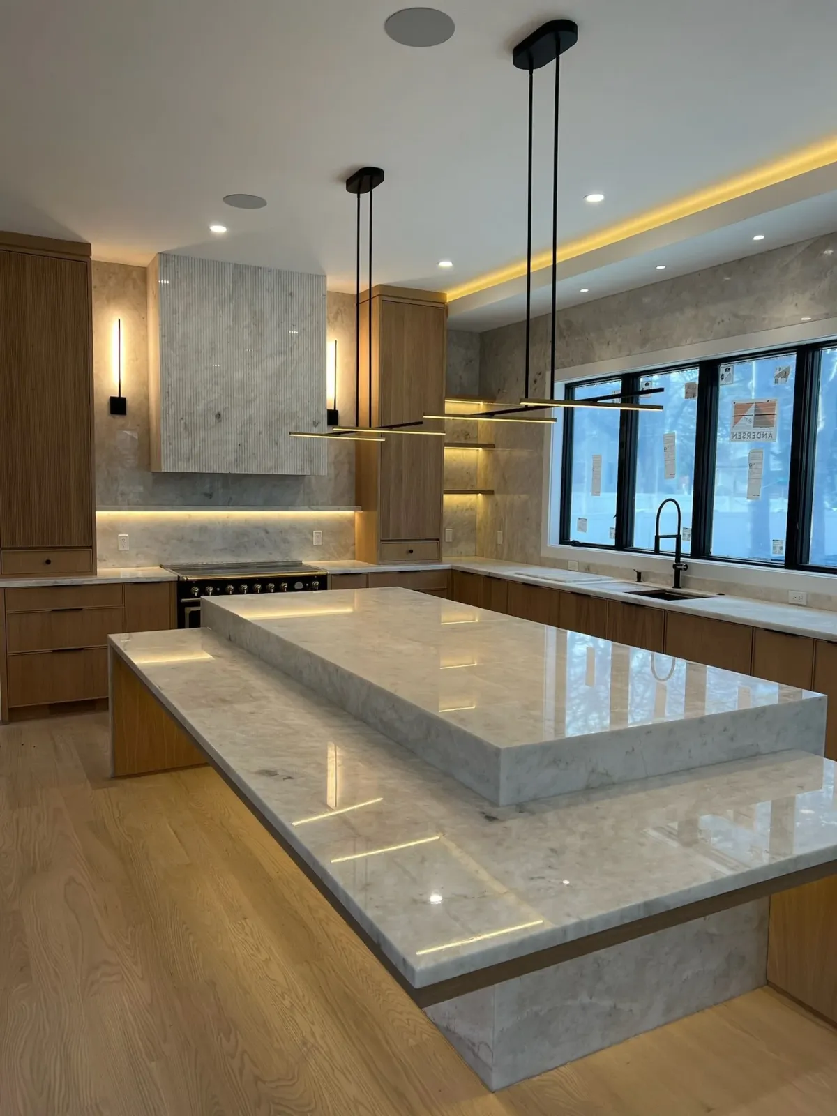 Modern kitchen with marble countertops, a large marble island, wooden cabinets, and a window overlooking outside. Pendant lights hang above the island, and there are LED strip lights built into the ceiling and behind the cabinets.