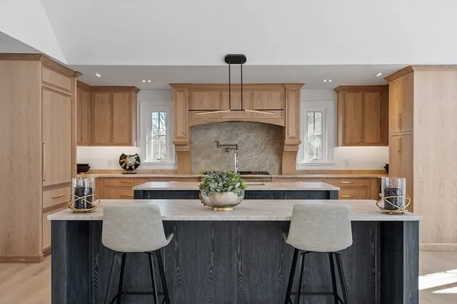 Modern kitchen with light wood cabinets, a marble island with two chairs, and a gray backsplash.