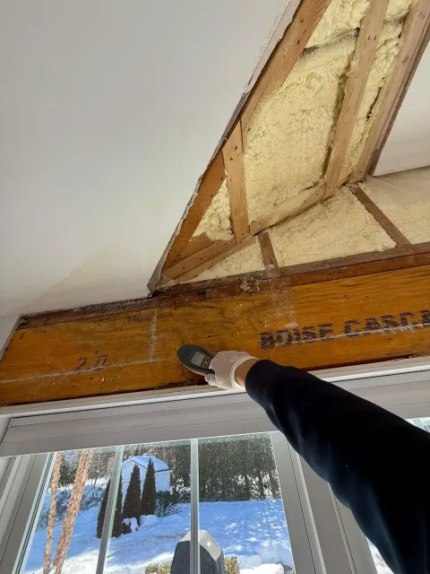Hand pointing at exposed wooden roof beam above a window in a snowy cabin interior
