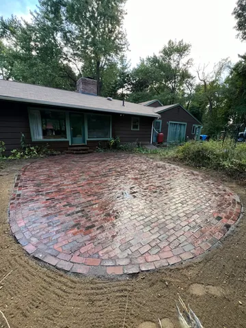A circular brick patio has been newly laid in a backyard, with the reddish-brown pavers arranged in a fan pattern radiating from the center. Fresh sand and unfinished landscaping surround the patio, while a covered porch and mature trees frame the outdoor space. Construction materials and tools in the background suggest the project is still in progress
