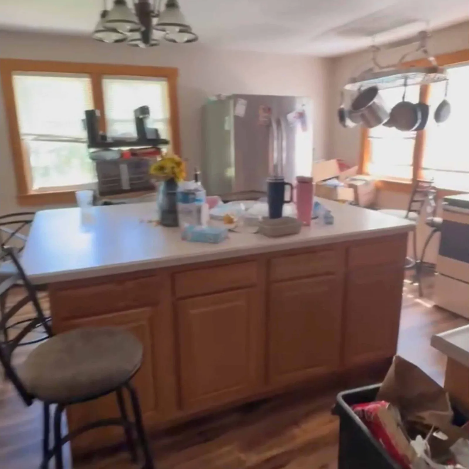Kitchen with wooden island cluttered with items, hanging pots, stools, and a stainless steel refrigerator.