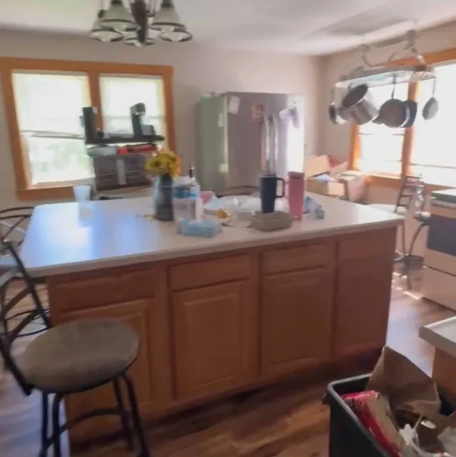 Kitchen with wooden island cluttered with items, hanging pots, stools, and a stainless steel refrigerator.