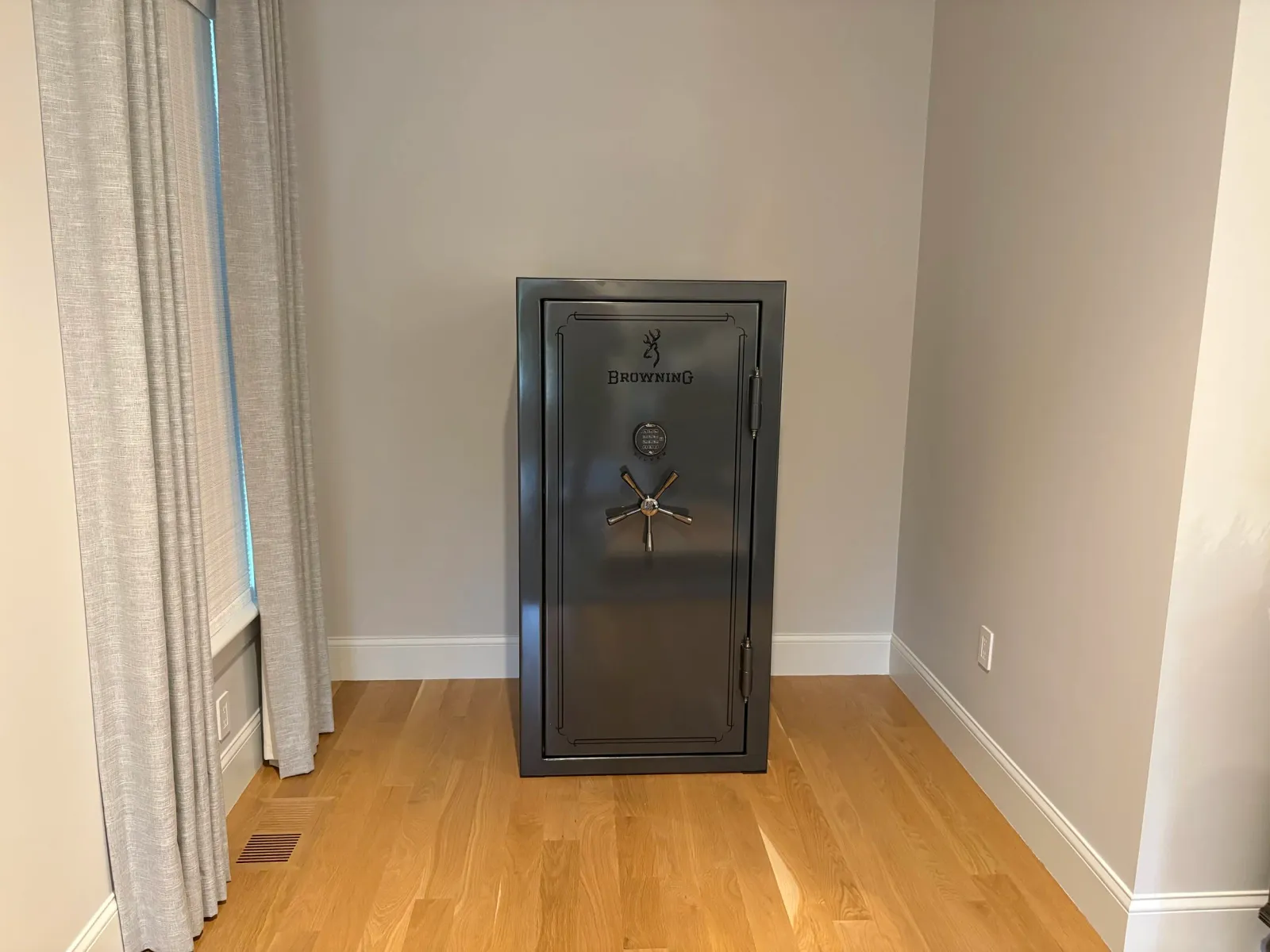 A gray Browning safe with a keypad lock and a handle, placed in the corner of a room with wooden flooring and cream-colored walls. There is a window with light-colored curtains on the left side of the image.