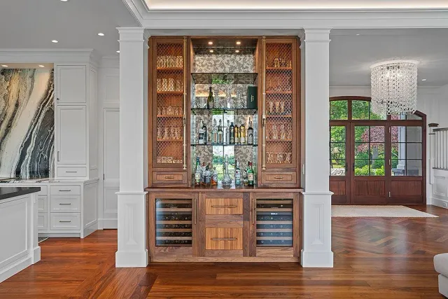 Wooden bar with wine fridges, shelves of liquor, and decorative glass.  Between white pillars in a room with hardwood floors.
