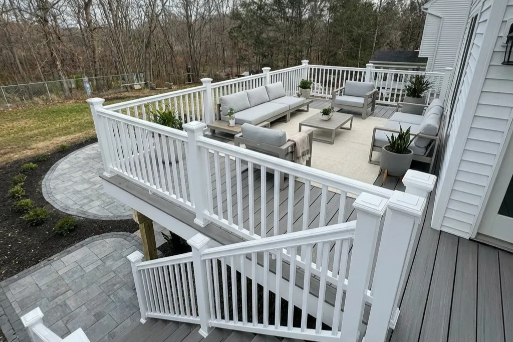 Outdoor upper deck patio with white railing, outdoor gray furniture including sofas and chairs, potted plants, and a stone pathway leading to a grassy yard surrounded by trees.