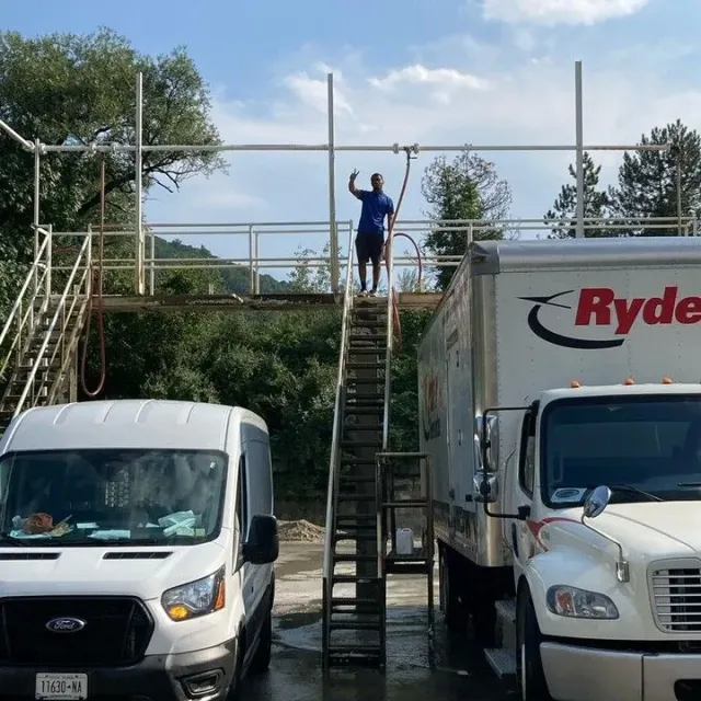 Man on a platform with a hose, giving a thumbs up.  Van and truck parked below, structure in a green setting.