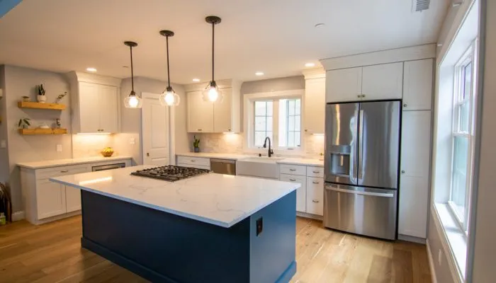 Spacious kitchen showcasing a striking blue island with white quartz countertop, three globe pendant lights, white cabinetry, stainless steel refrigerator, farmhouse sink, and natural light from oversized windows.