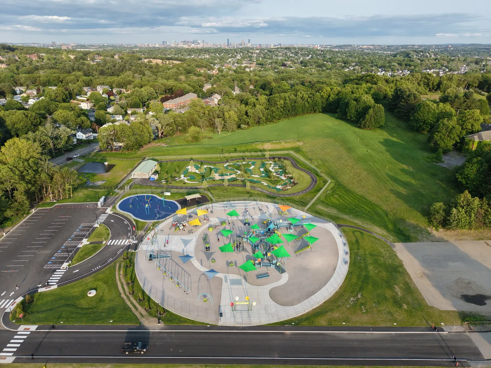 Aerial view of a park with a colorful playground, water features, mini golf course, parking lot, sports field, green trees, and surrounding residential area with a city skyline in the distance.