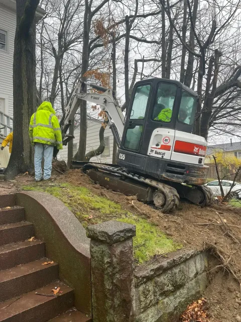 Worker beside mini excavator on a muddy slope next to a house during outdoor maintenance work