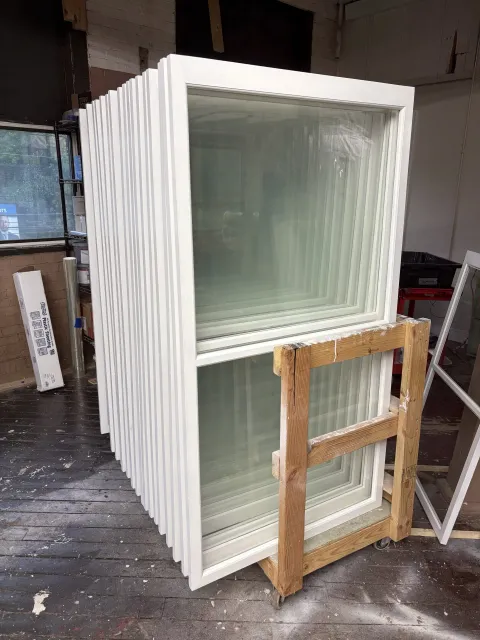 Stack of white window frames leaning against a wooden cart in a workshop or warehouse setting.