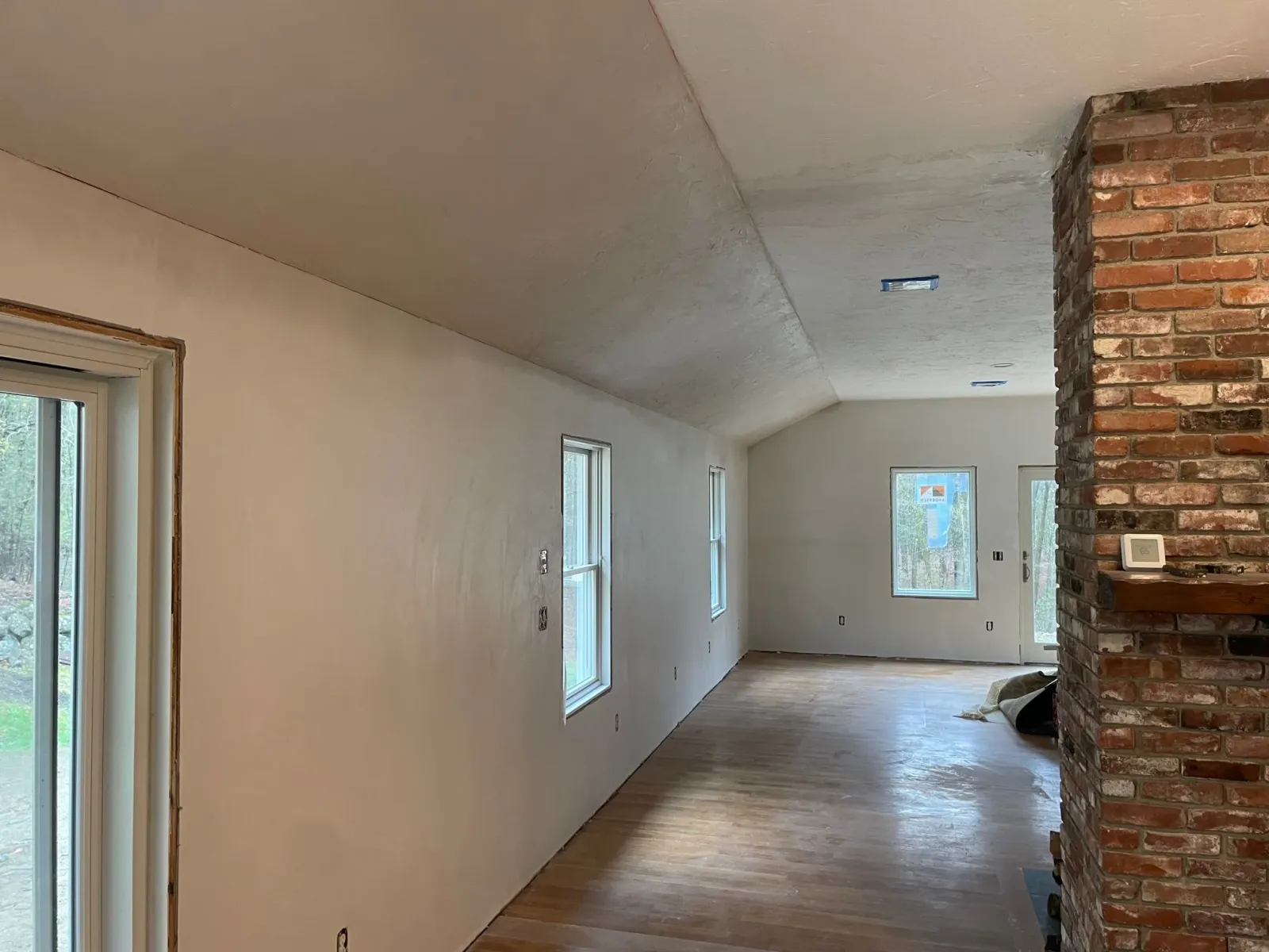 Interior of a house under construction with white finished walls, wooden floor, multiple windows, a brick pillar, and a temporary thermostat on the brick wall.