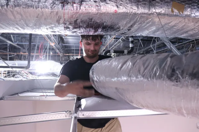A young man with wet hair working underneath a large metallic pipe in an industrial or manufacturing setting, with exposed pipes and wiring above him.