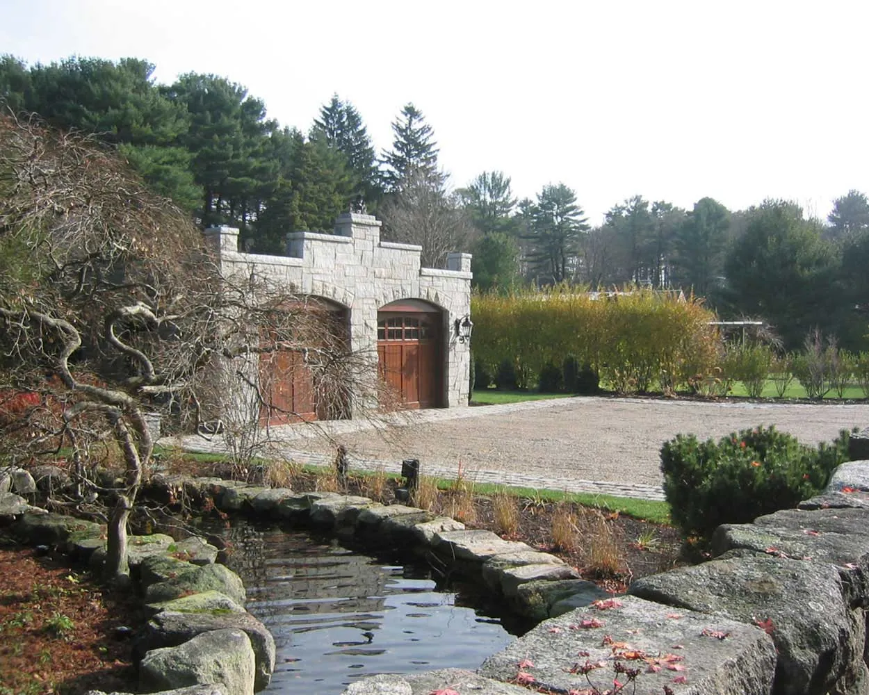 Stone carriage house with arched wooden doors, set beside a gravel driveway and a landscaped pond edged with large rocks and ornamental trees.