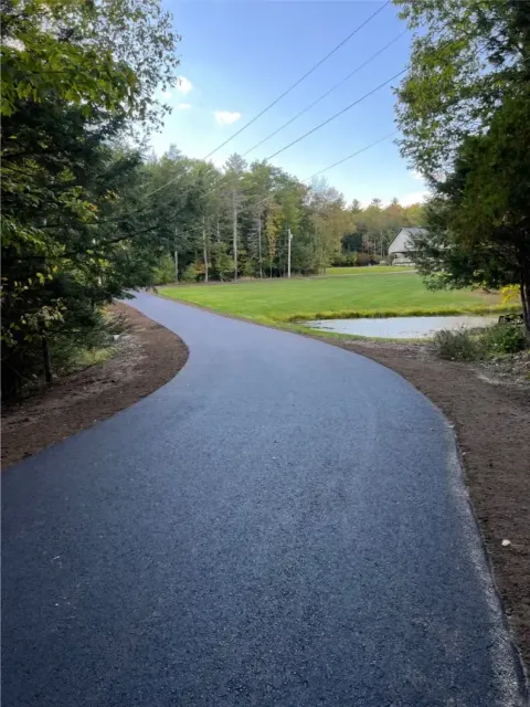 Paved road curves through a green, wooded area.  A grassy lawn with a pond and house are in the distance.