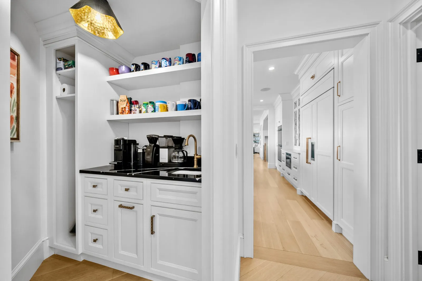 Custom butler's pantry with white cabinetry, brass hardware, black countertops, and a built-in coffee station, opening to a hallway with floor-to-ceiling white built-ins and hardwood floors