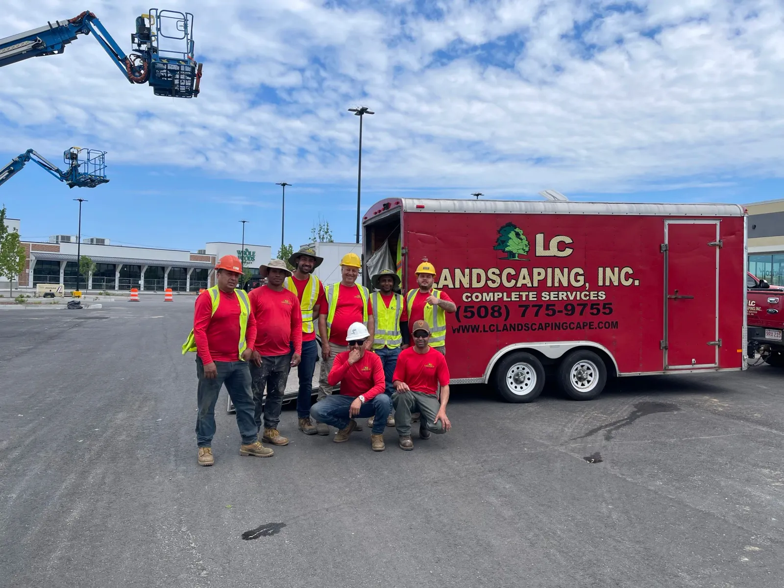 Group of workers posing in front of red LC Landscaping, Inc. trailer at a parking lot with construction equipment in the background.