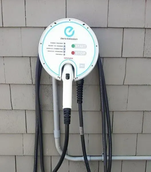 Electric vehicle charger mounted on a gray, wooden building.  White and blue circular charging station with two cables.