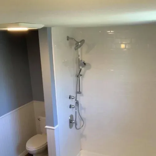 A bathroom features a white shower with multiple showerheads and controls, set against a tiled wall. A toilet is visible in the corner next to a gray and white half-paneled wall.