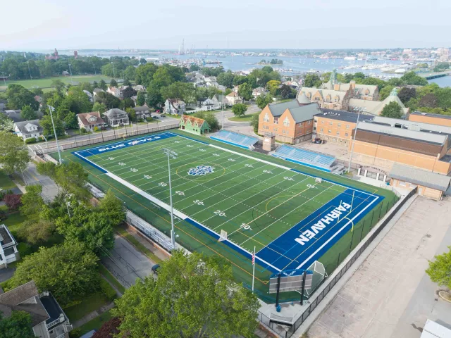 Aerial view of a football field surrounded by trees and houses, with water and boats in the background. The field has blue end zones labeled "KAHUEN" and "BLUE FALCONS".