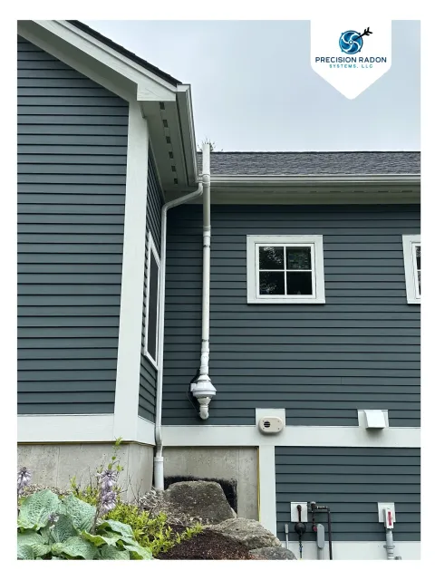 Exterior view of a house with dark blue horizontal siding, white trim, and a window, showing a radon vent mitigation system removing radon gas.